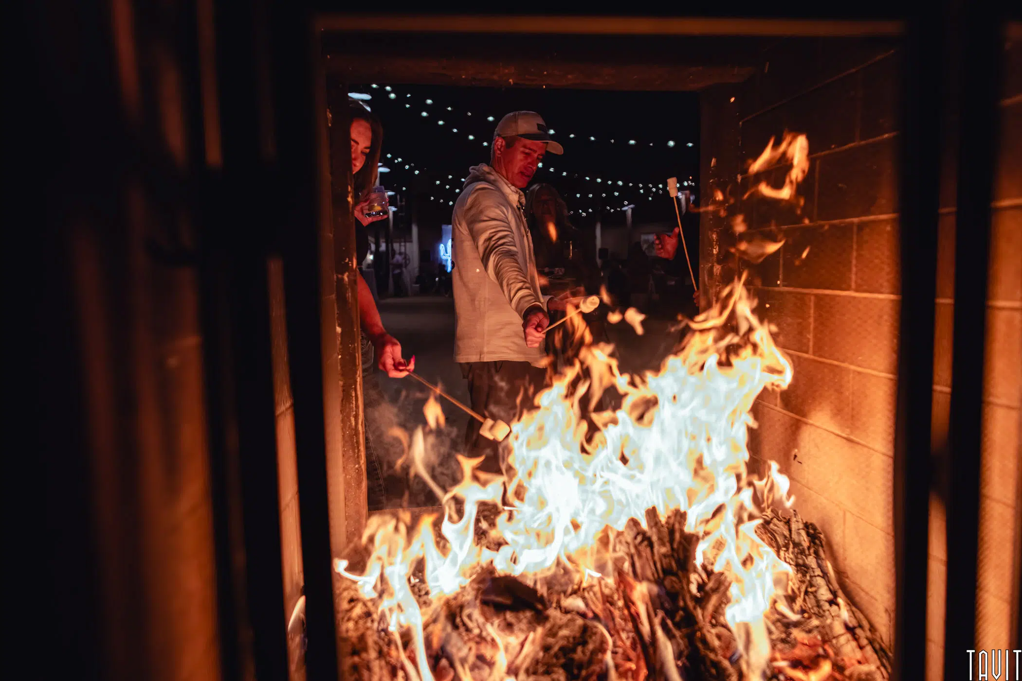 A man in a light hoodie and cap stands by an outdoor fire pit at night, holding a stick over the flames. String lights and another person are visible in the background.