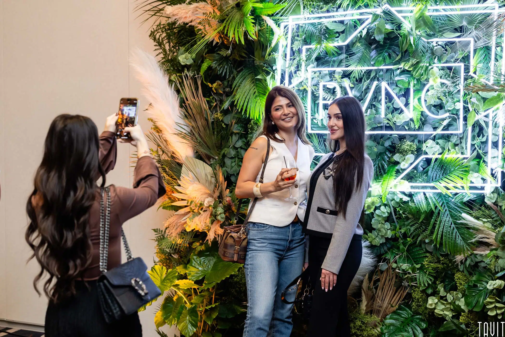 Two women pose and smile for a photo in front of a neon “SYNC” sign with lush greenery. Another woman takes their picture with a smartphone. All are dressed in business casual.