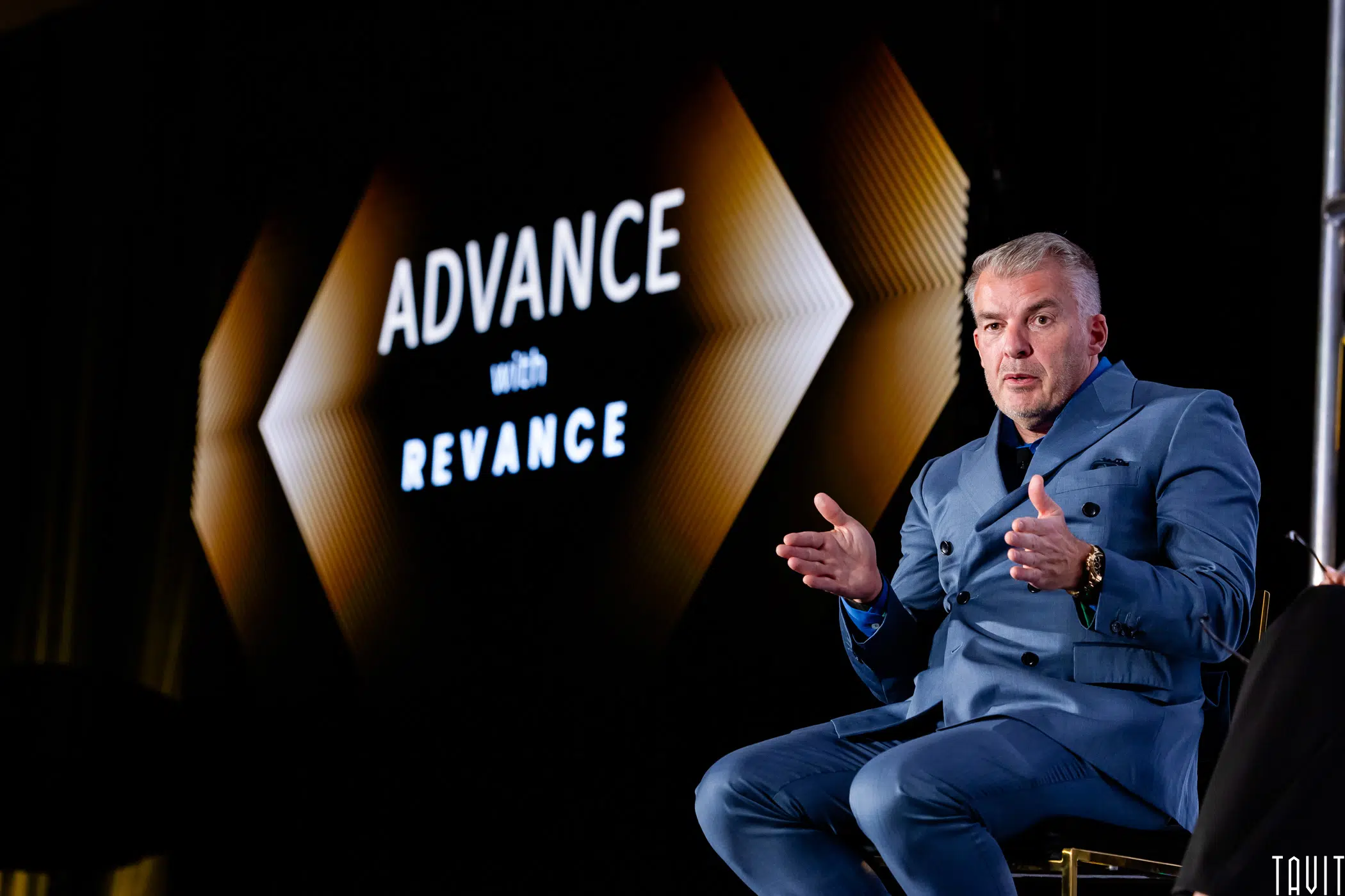 A man in a blue suit gestures while speaking on stage in front of a black backdrop with the words “ADVANCE with REVANCE” in white text.