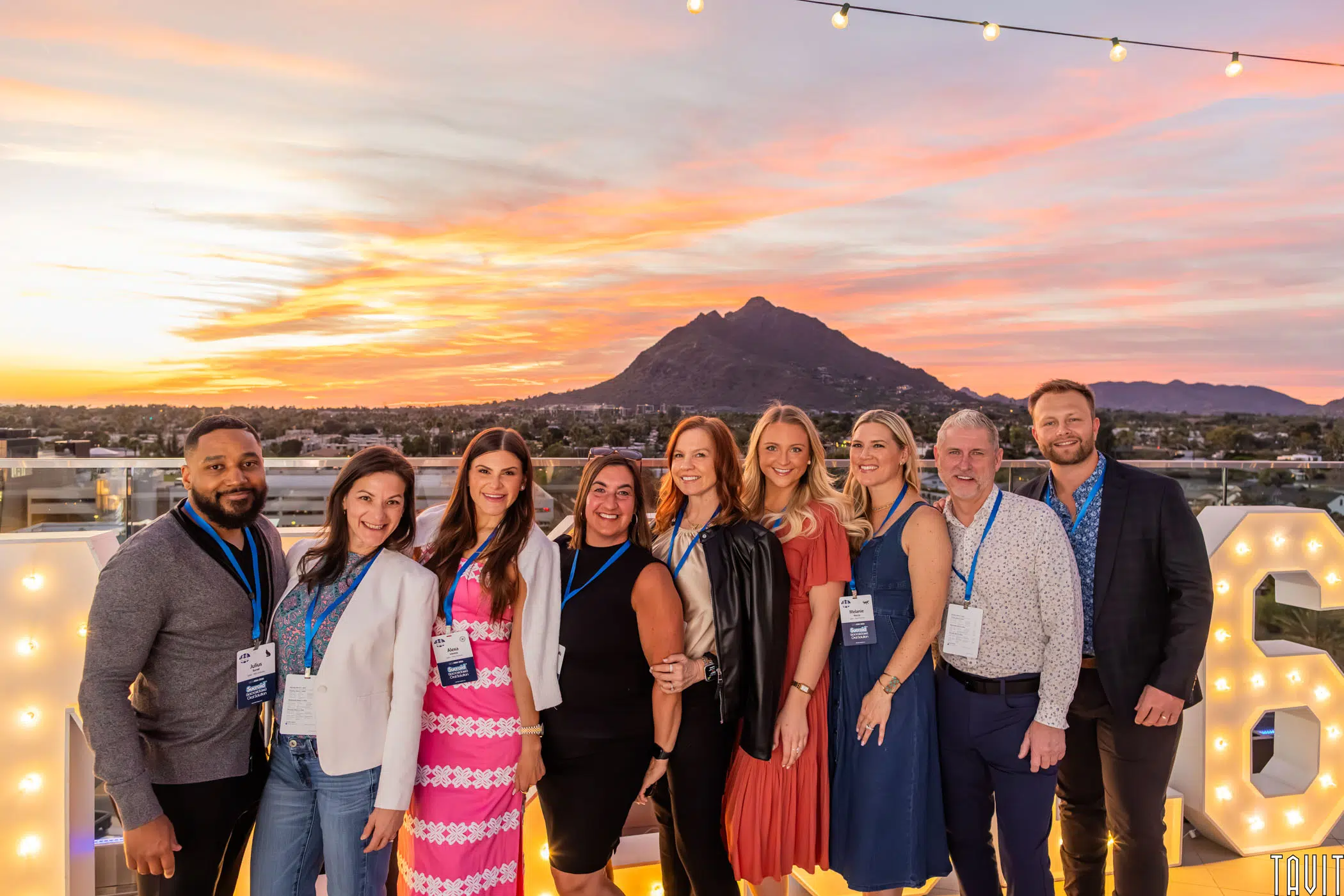 Nine people pose and smile on a rooftop at sunset, with a mountain and city in the background. They wear conference badges and business casual to semi-formal attire. String lights are overhead, and large lit numbers are behind them.
