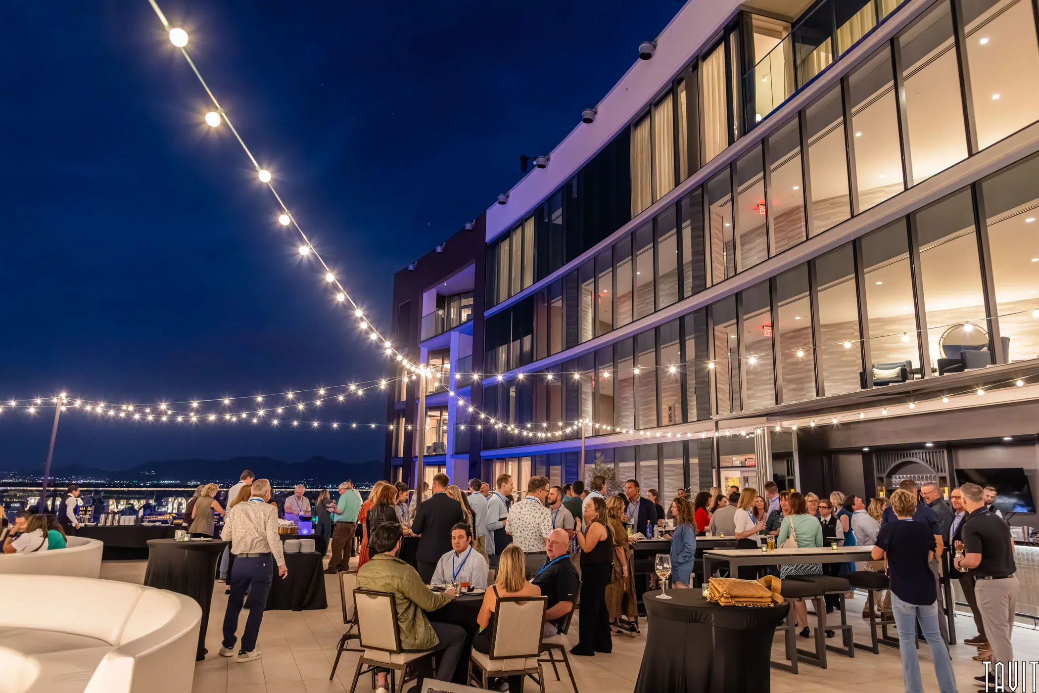 A lively rooftop party at night features people mingling under string lights, with a modern glass building in the background and city lights visible in the distance. Tables and chairs are arranged across the patio.