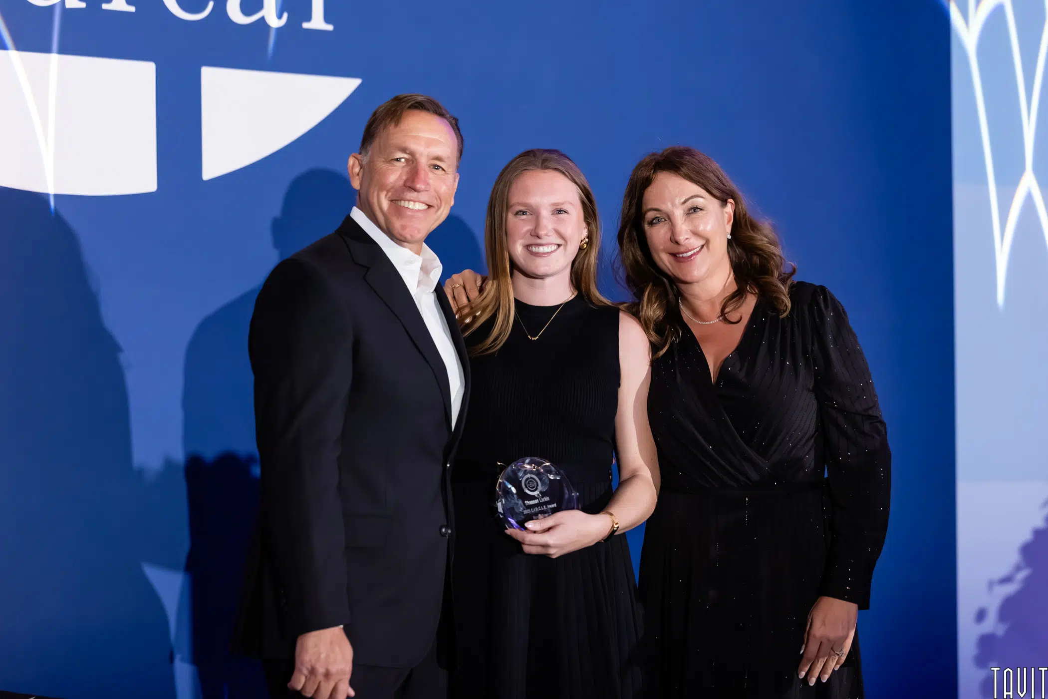 Three people in formal attire stand together at an awards event. The woman in the middle is holding a glass award and smiling, with two people standing close on each side against a blue backdrop.