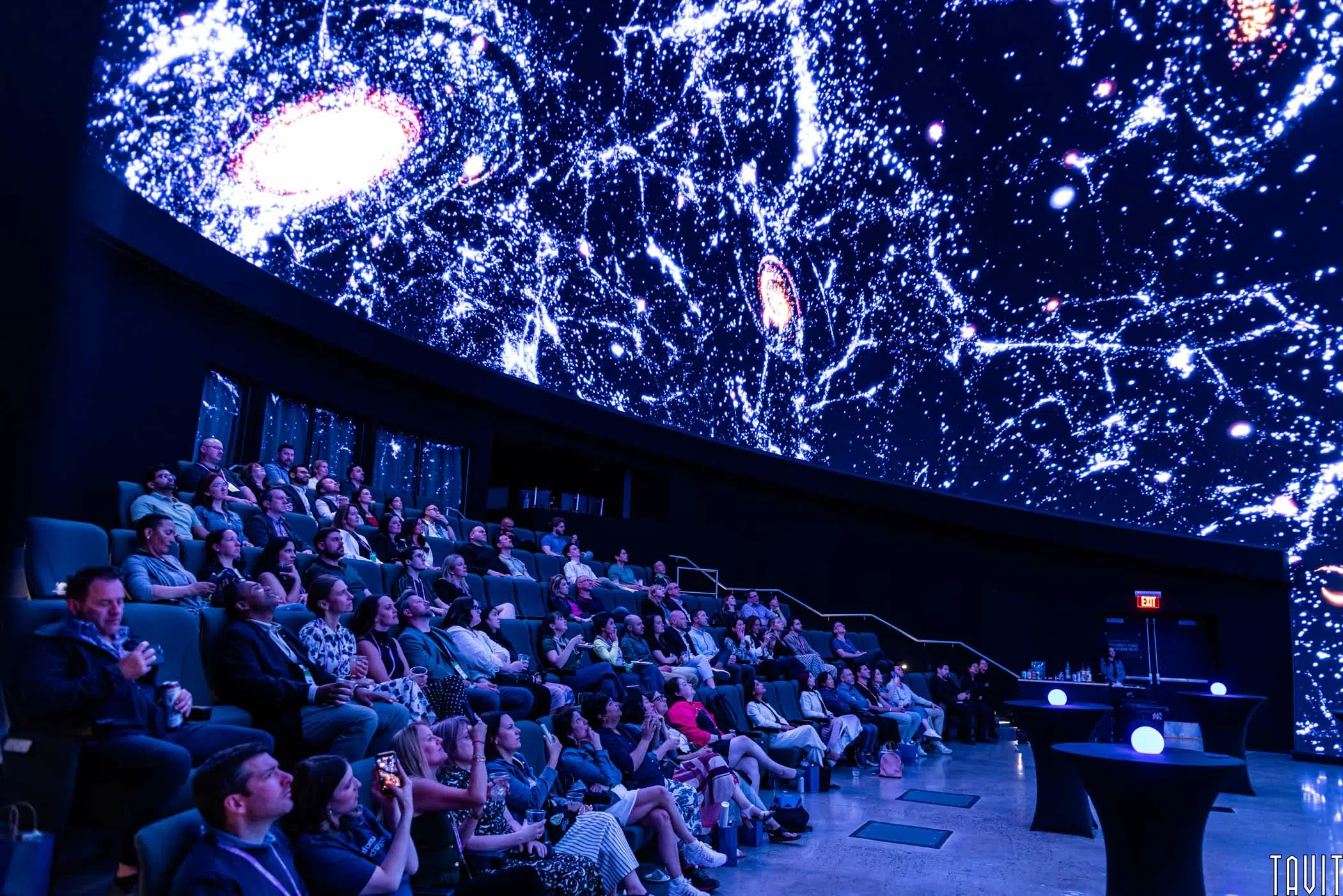 Audience seated in a dark planetarium, watching a dome-shaped screen displaying colorful galaxies and stars in space.