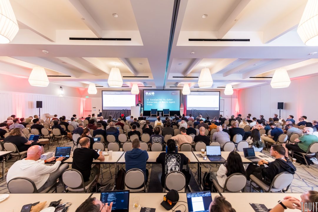 A large group of people attend the OAIC conference in a bright, modern hall, facing a stage with three projection screens displaying presentation slides.