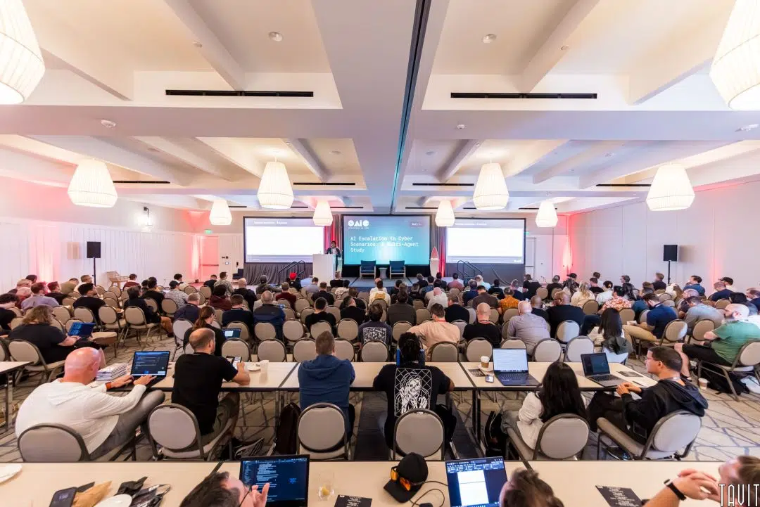 A large group of people attend the OAIC conference in a bright, modern hall, facing a stage with three projection screens displaying presentation slides.