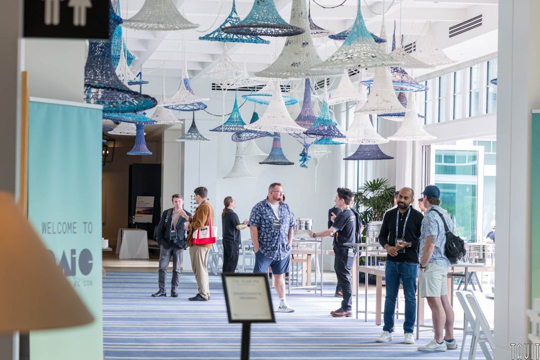 OAIC attendees stand and chat in a bright, modern lobby with large windows and hanging woven sculptures. Some hold bags or wear badges, suggesting an event or conference setting. Tables and chairs are scattered around the space.