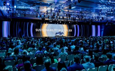 A large audience sits in a dimly lit conference hall at a corporate event, facing a stage with the words "SUSTAINABILITY MULTIPLIER" projected on a screen, surrounded by blue lighting and decorative ceiling lights.