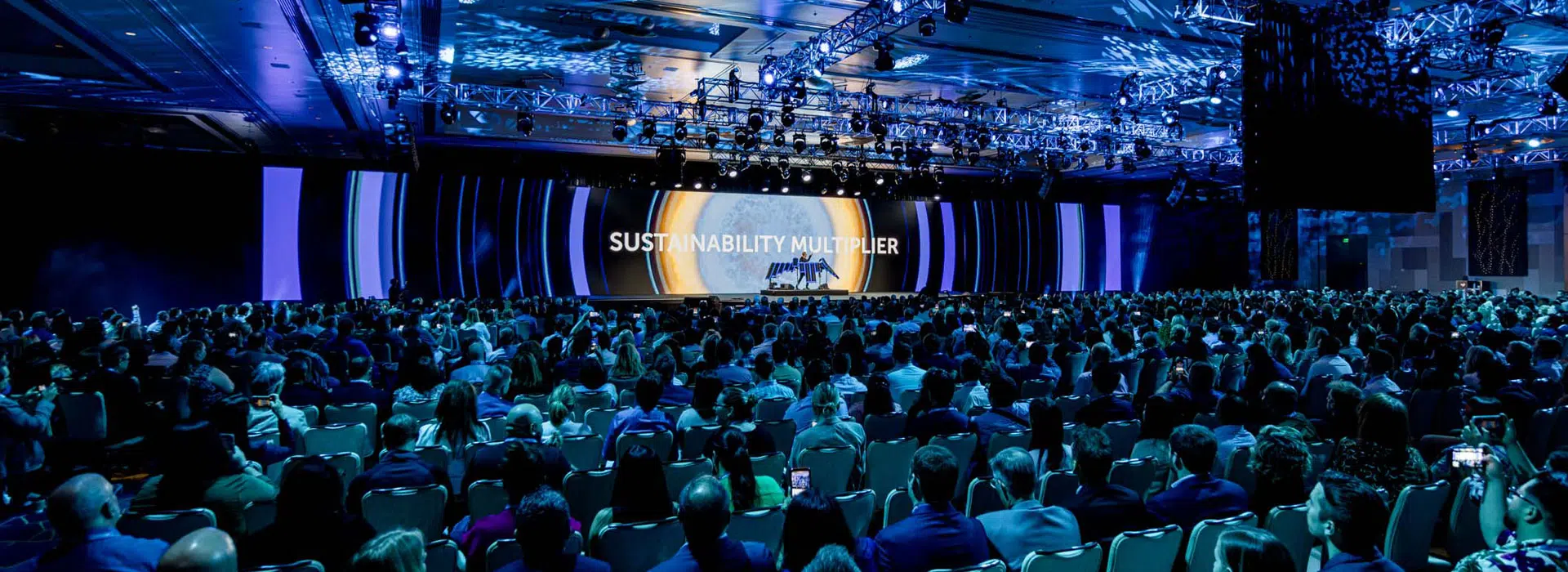 A large audience sits in a dimly lit conference hall at a corporate event, facing a stage with the words "SUSTAINABILITY MULTIPLIER" projected on a screen, surrounded by blue lighting and decorative ceiling lights.