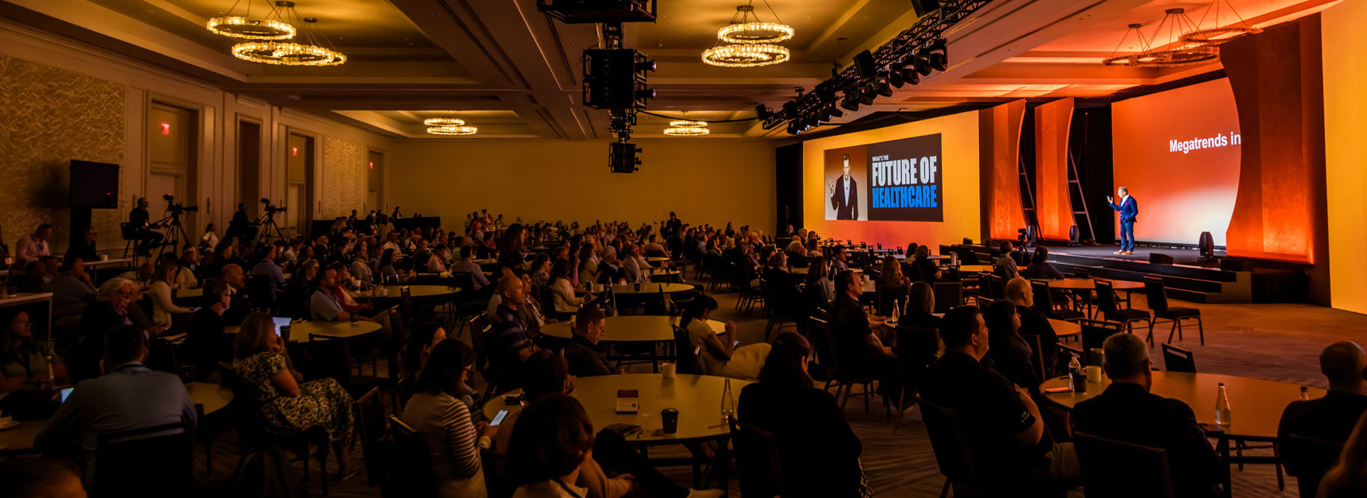 A large audience sits at round tables in a dimly lit conference room, watching a speaker on stage with a screen displaying "The Future of Healthcare." Warm lights illuminate the stage area.