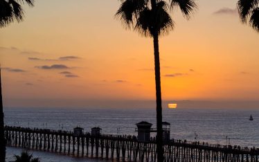 Tall palm trees frame a pier in San Diego, extending into the ocean at sunset, with orange and purple hues in the sky and gentle waves on the water.