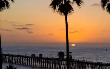 Tall palm trees frame a pier in San Diego, extending into the ocean at sunset, with orange and purple hues in the sky and gentle waves on the water.