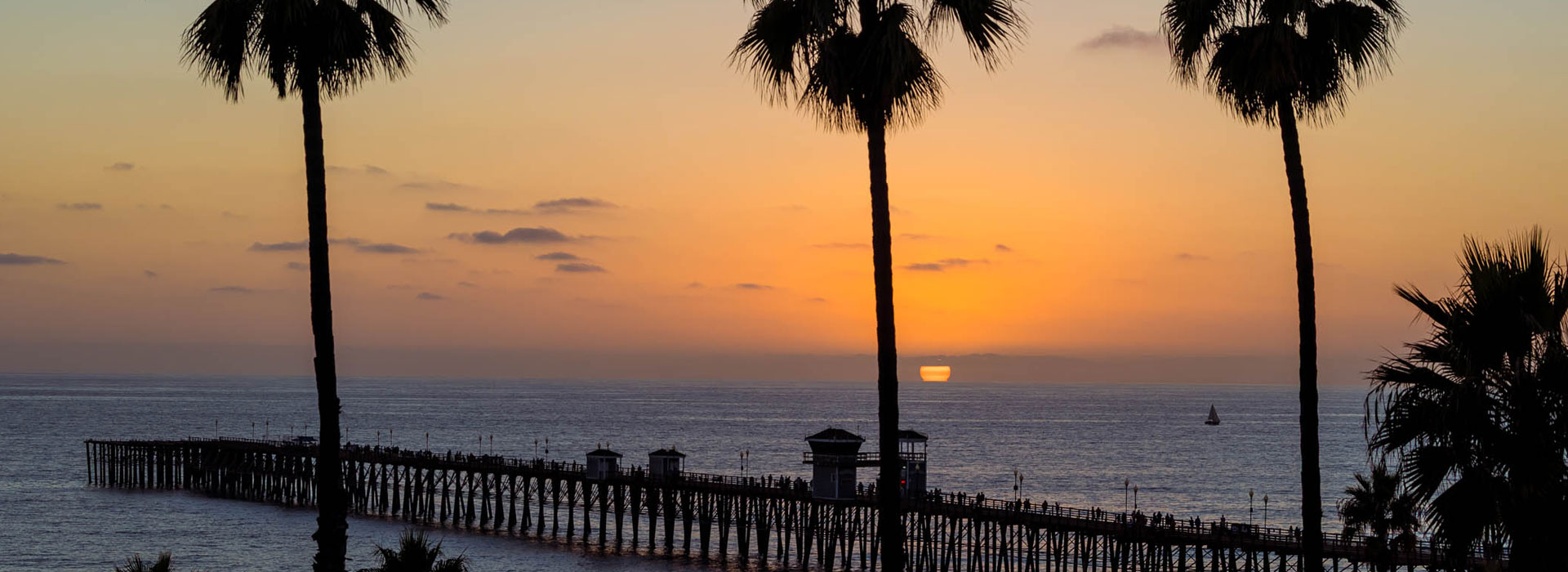 Tall palm trees frame a pier in San Diego, extending into the ocean at sunset, with orange and purple hues in the sky and gentle waves on the water.