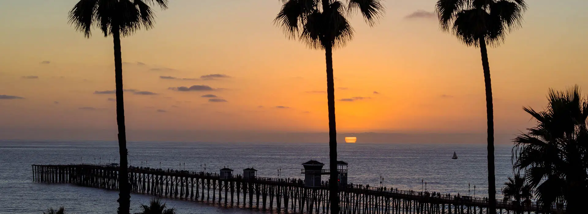 Tall palm trees frame a pier in San Diego, extending into the ocean at sunset, with orange and purple hues in the sky and gentle waves on the water.
