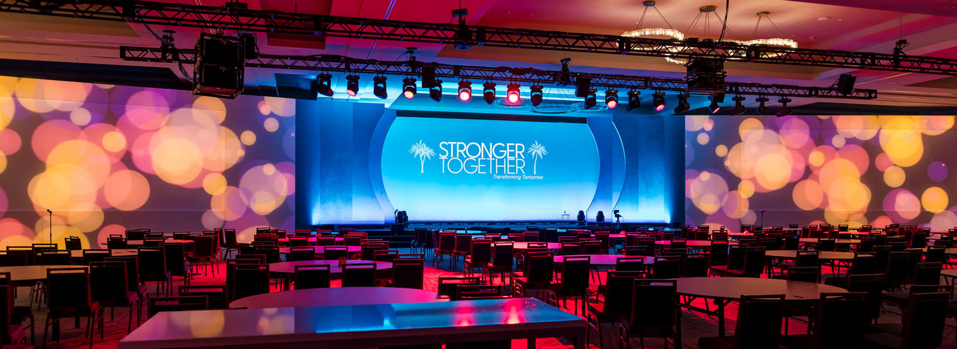 Large conference room with rows of chairs and tables facing a brightly lit stage. The backdrop reads "Stronger Together" with colorful circular light patterns projected across the walls, perfectly captured by a professional Scottsdale event photographer.
